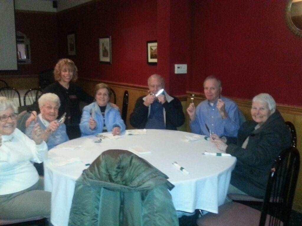 A group of people sit around a circular table in a restaurant with red walls, smiling and holding silverware.