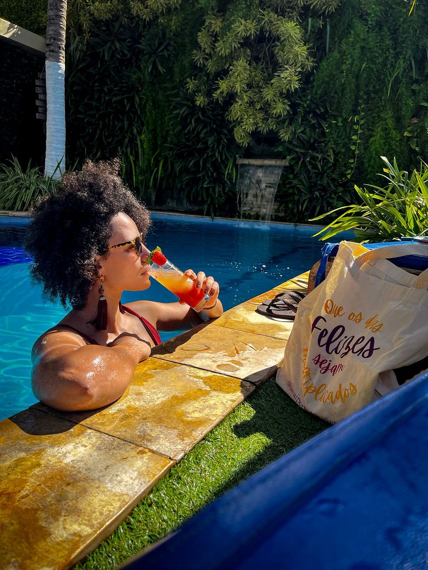 Mulher relaxando em uma piscina, tomando um drinque ao lado de uma bolsa no deck, com vegetação exuberante ao fundo.