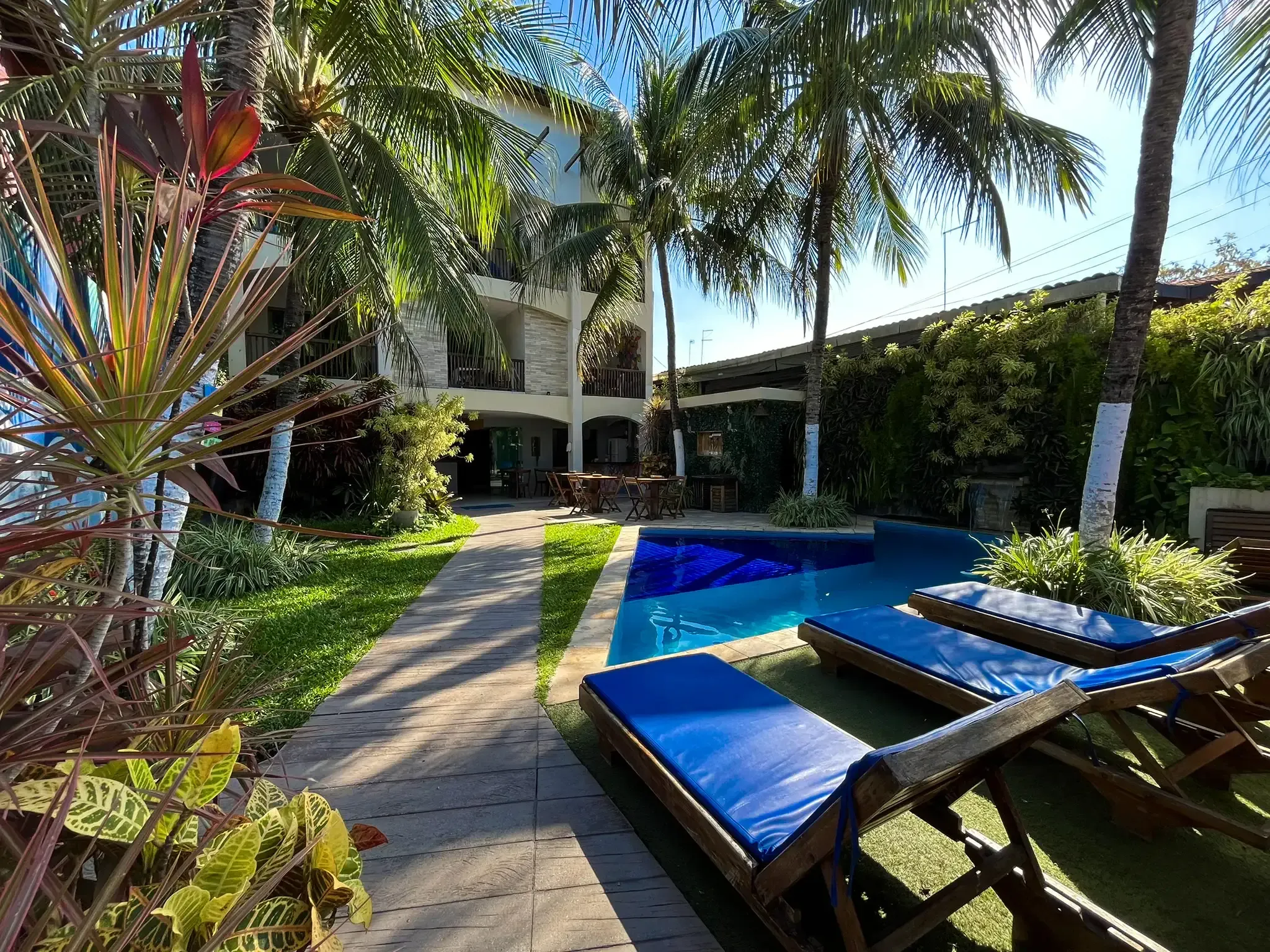 Pátio tropical à beira da piscina com espreguiçadeiras azuis, palmeiras e uma passarela ao lado de um edifício moderno.