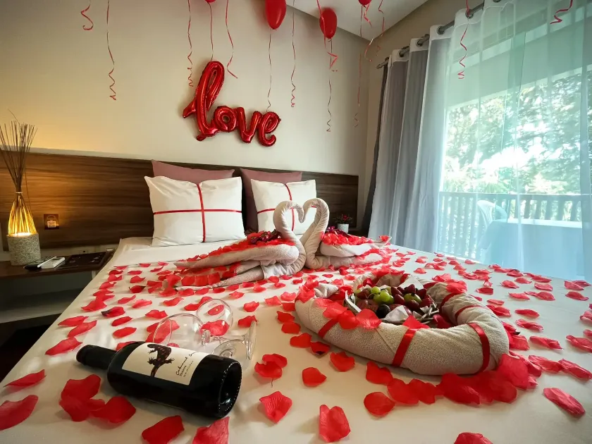 Cama de hotel romântica com pétalas de rosa, decoração em forma de coração, garrafa de vinho e toalha em formato de cisne.