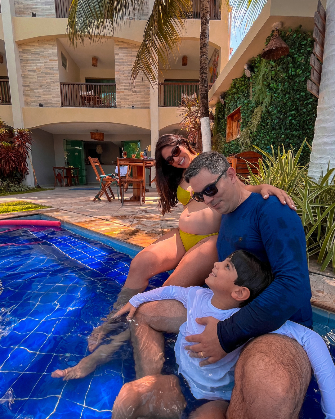 Homem e criança sentados à beira de uma piscina azul, com uma mulher atrás deles, em um pátio ensolarado.