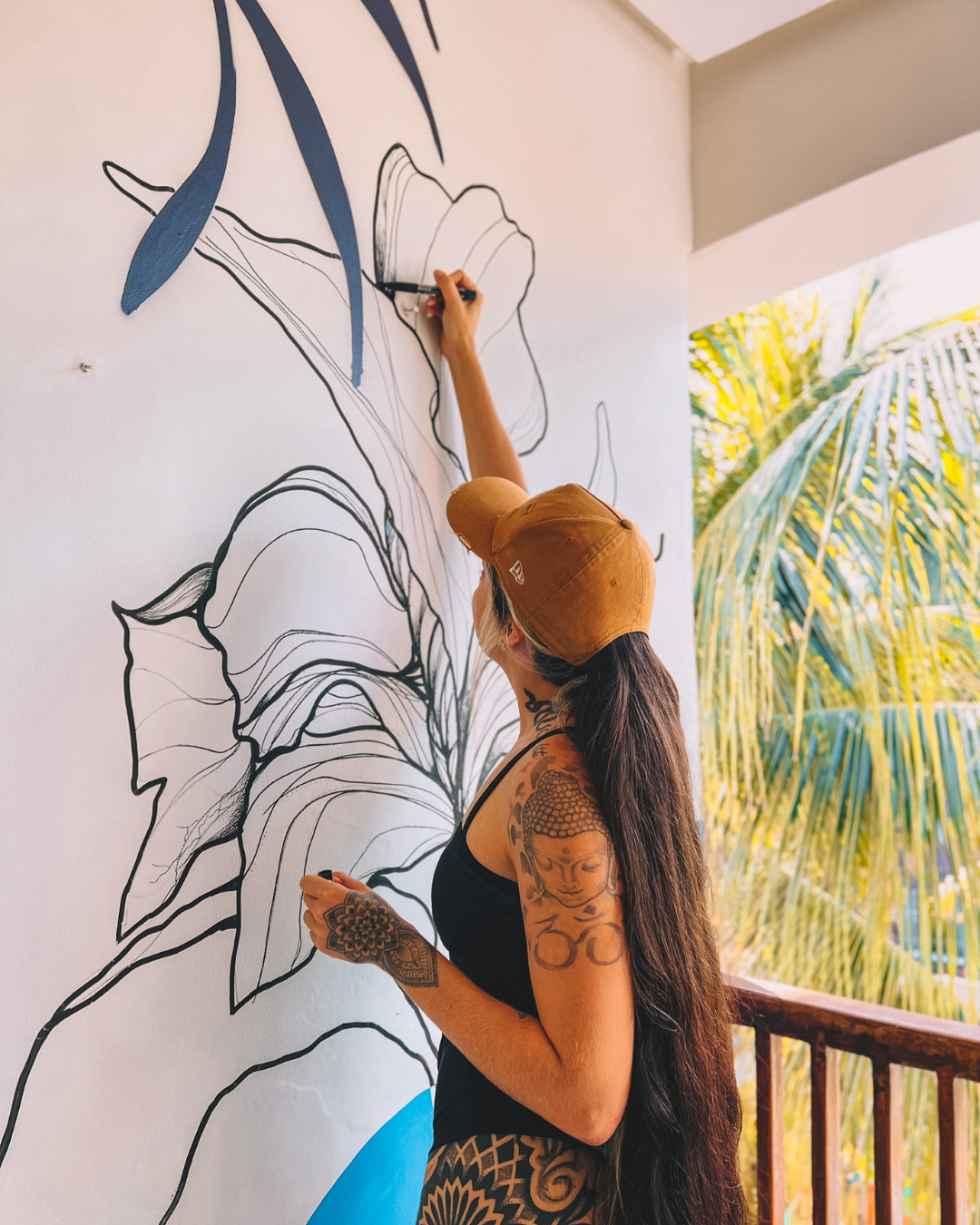 Pessoa pintando um mural em uma parede branca ao lado de uma varanda com palmeiras visíveis do lado de fora.