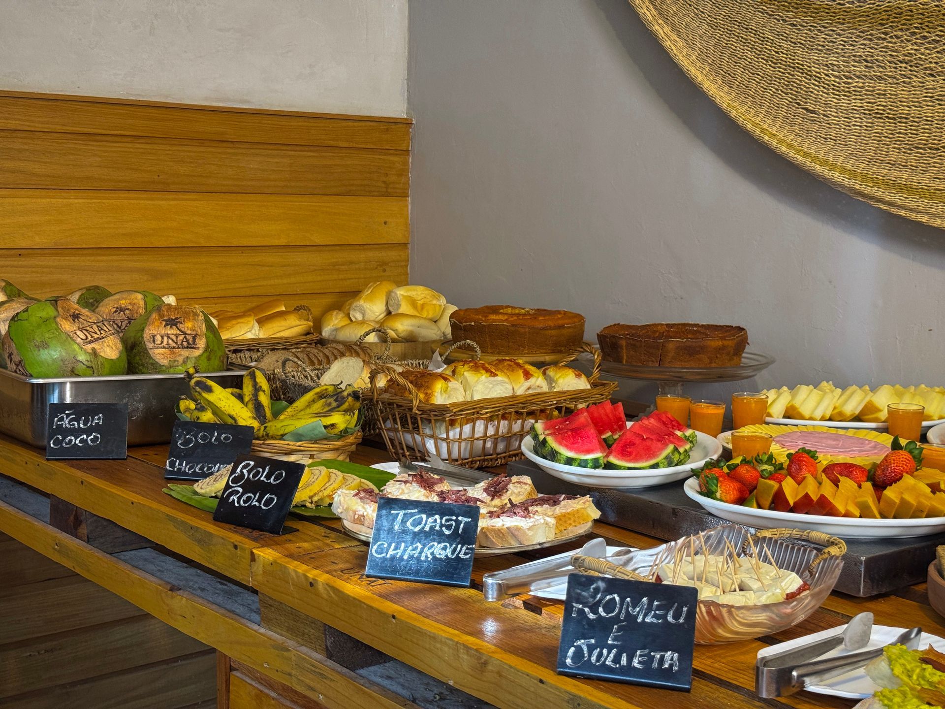 Mesa de bufê com uma variedade de doces, frutas e bandejas de sobremesas, todas identificadas com pequenas placas de giz.