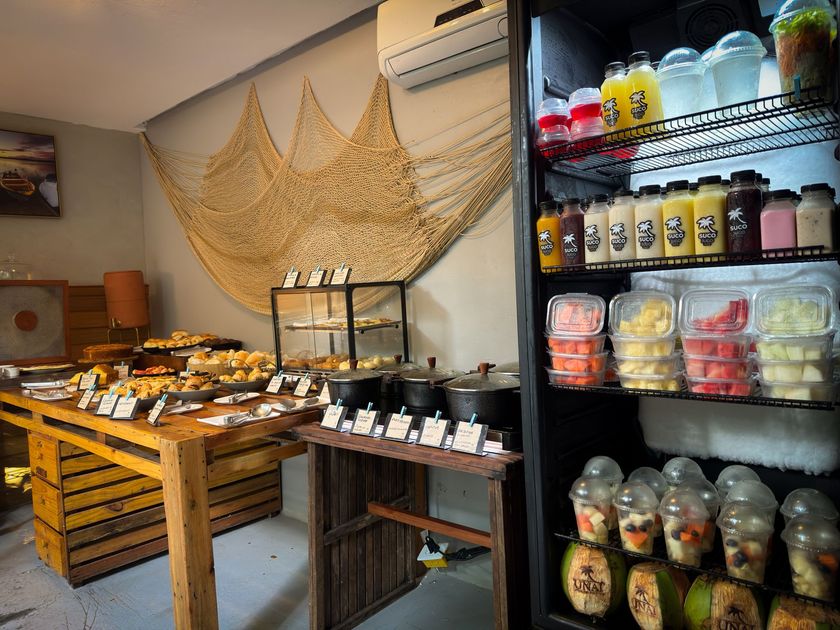Vitrine de padaria com doces em mesas de madeira e prateleira refrigerada com bebidas e alimentos embalados.