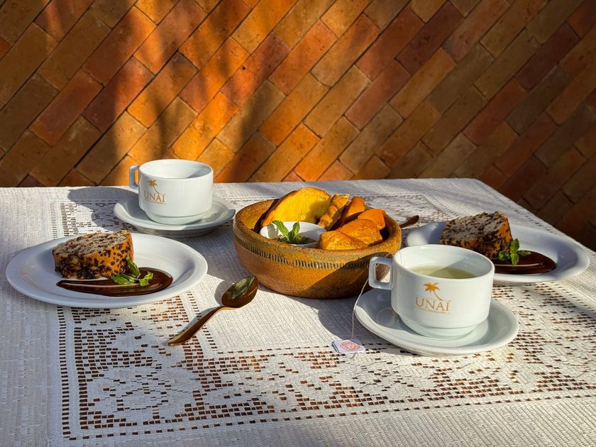 Mesa de chá ao ar livre com xícaras, doces e frutas, banhada de sol, encostada em uma parede de tijolos.