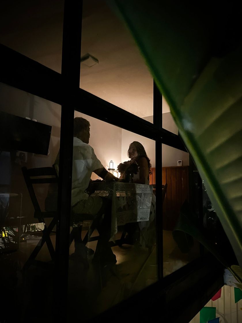 Duas pessoas conversando à mesa à luz de velas, vistas através de molduras escuras de janelas à noite.