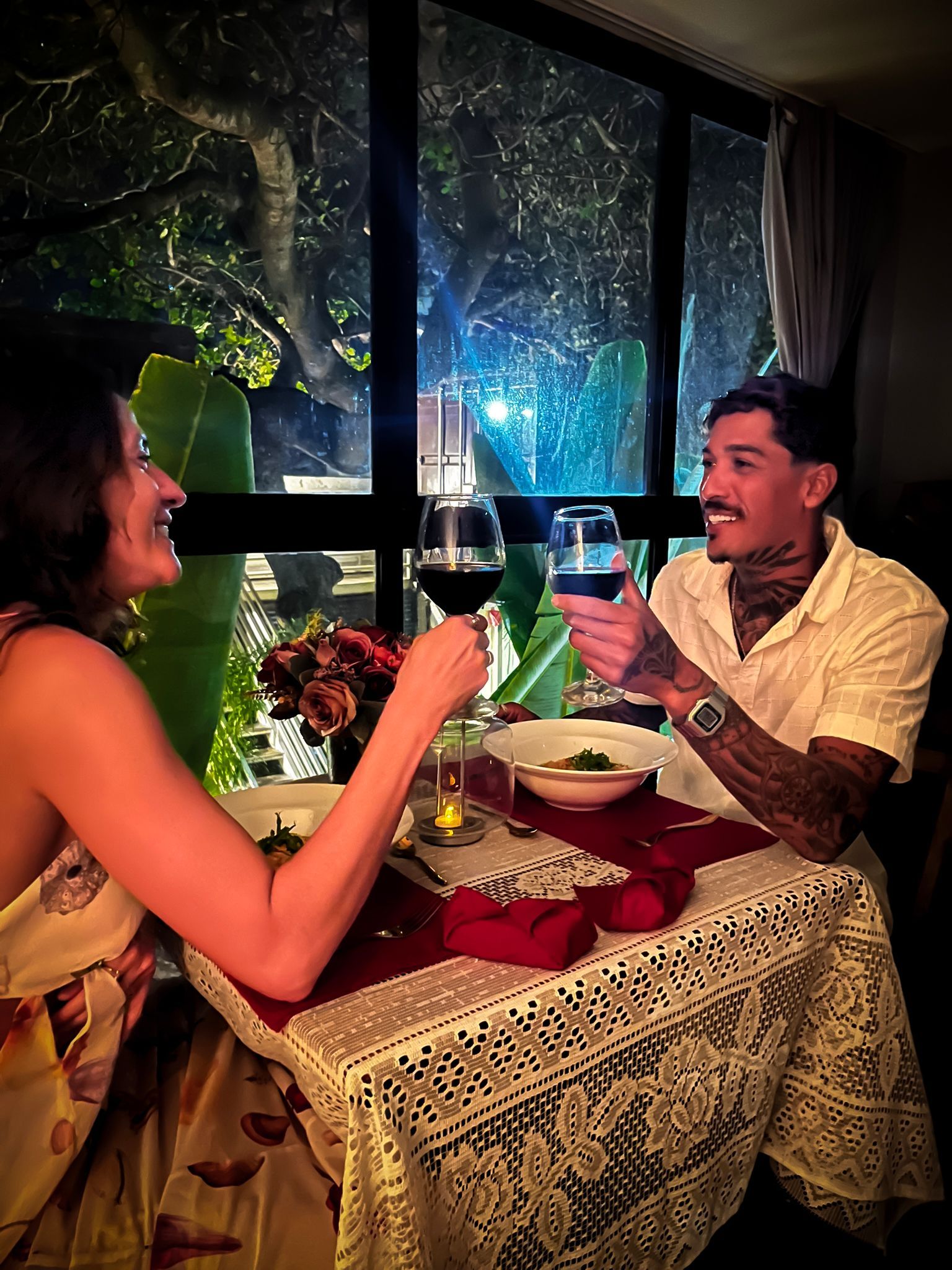 Duas pessoas brindam com vinho à luz de velas, junto a uma janela salpicada de chuva, à noite.