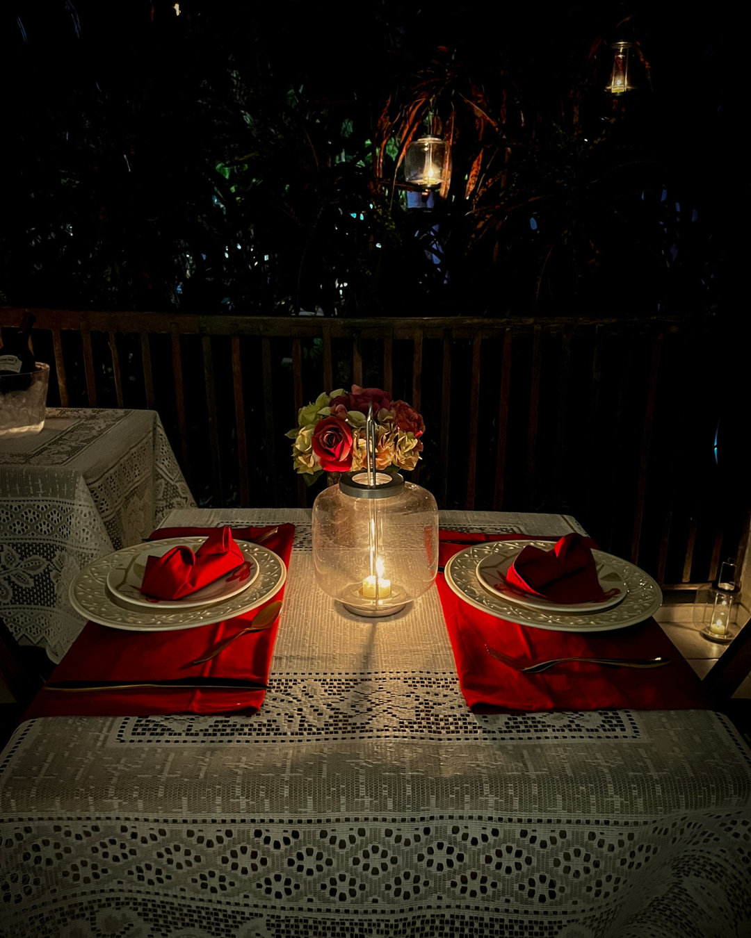 Mesa romântica à luz de velas em um deck com guardanapos vermelhos, flores e taças de cristal à noite.