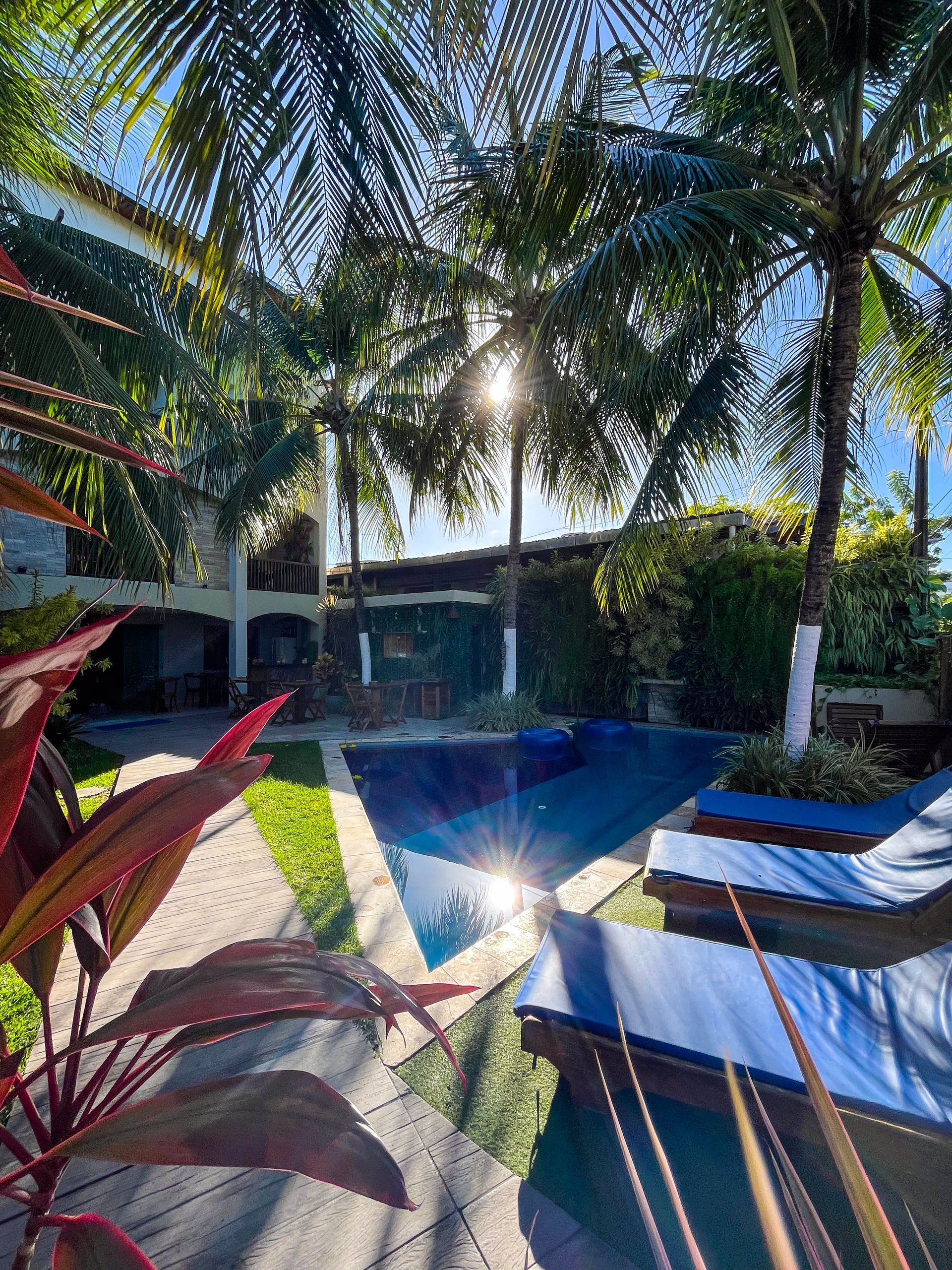 Área de estar tropical à beira da piscina, com palmeiras, água azul e espreguiçadeiras ensolaradas no pátio de um resort.