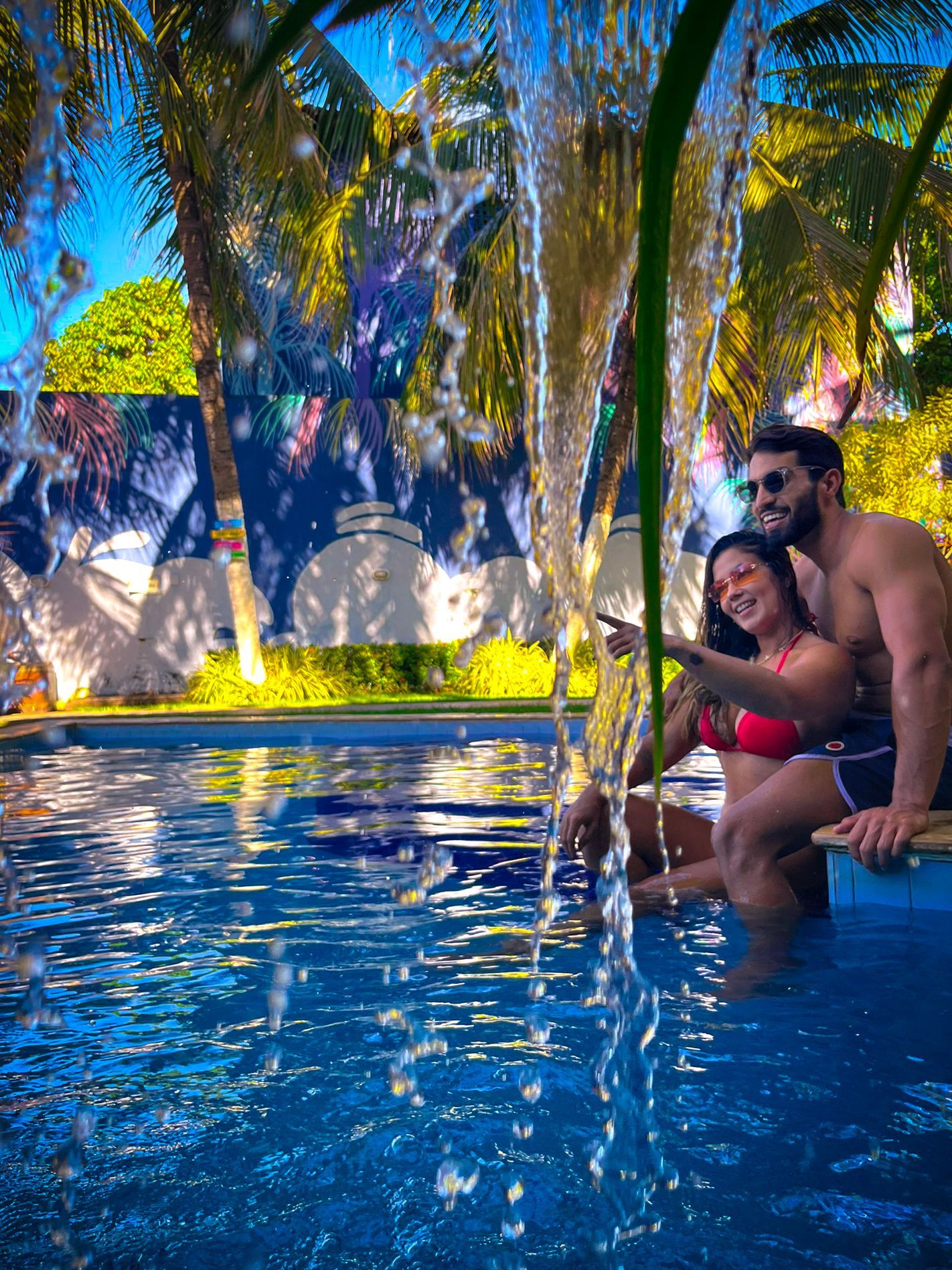 Casal sentado à beira de uma piscina com jatos de água, palmeiras e um cenário tropical vibrante.