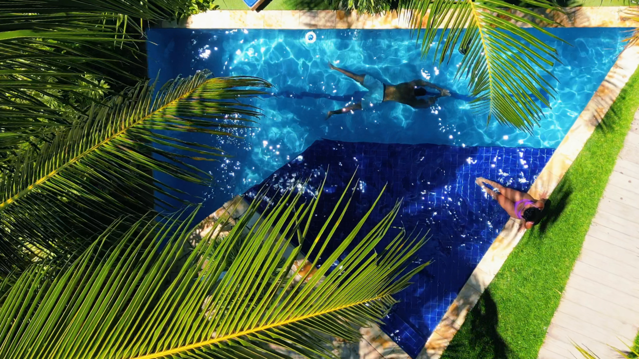 Vista aérea de uma piscina azul rodeada por palmeiras e uma passarela, com uma pessoa relaxando na borda.
