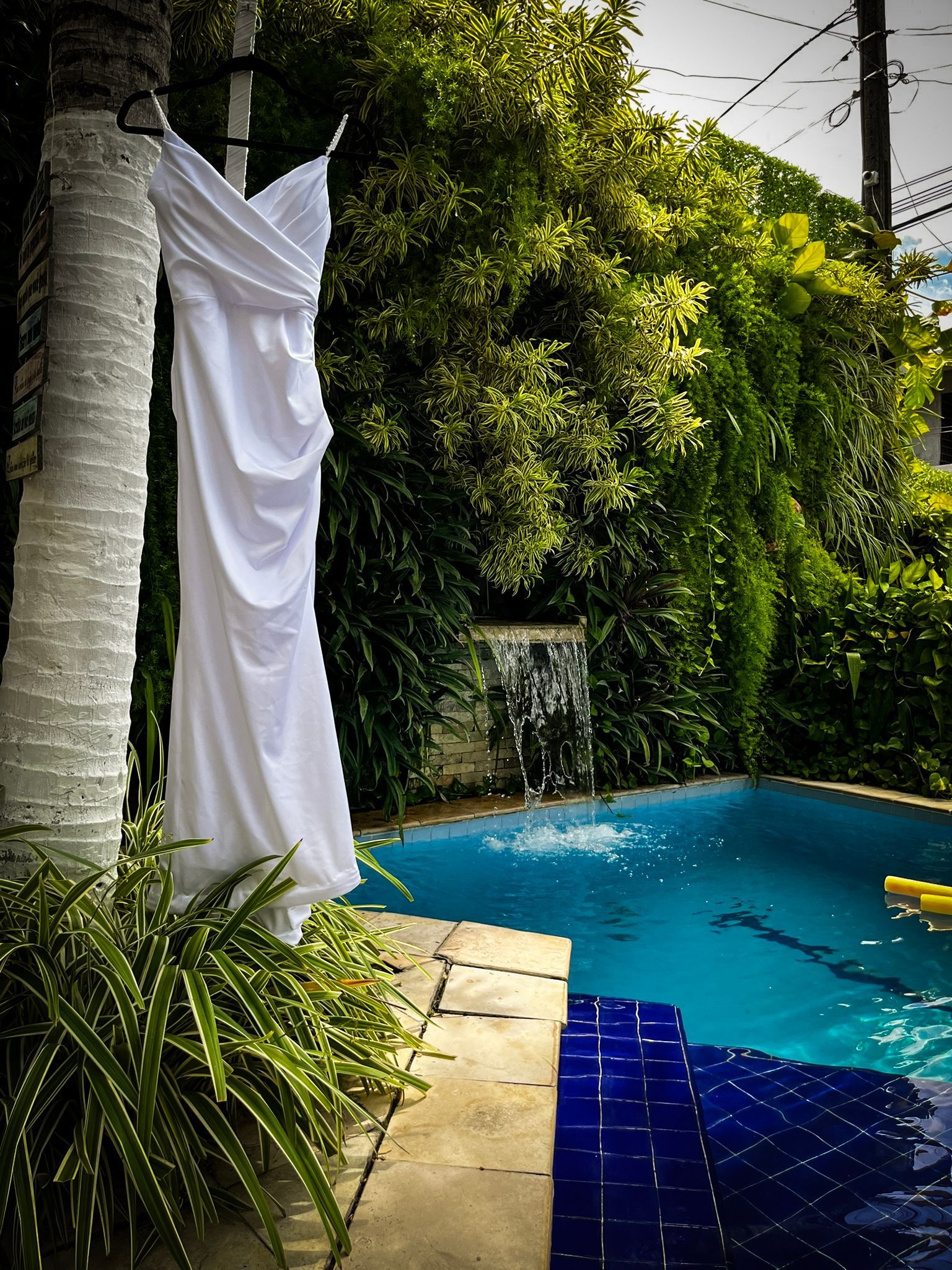 Cortina branca pendurada junto a uma piscina exuberante em um jardim com água azul e deck de azulejos.