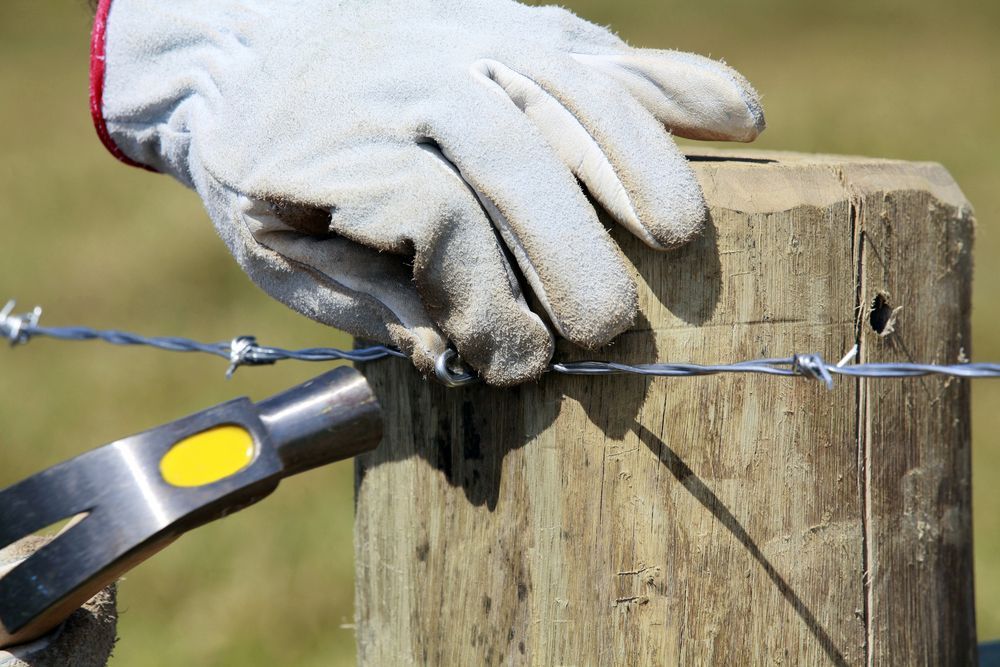 Hand Wearing Glove Securing Barbed Wire to Wooden Fence Post With a Hammer — Slash Mulch Co in Googa Creek, QLD