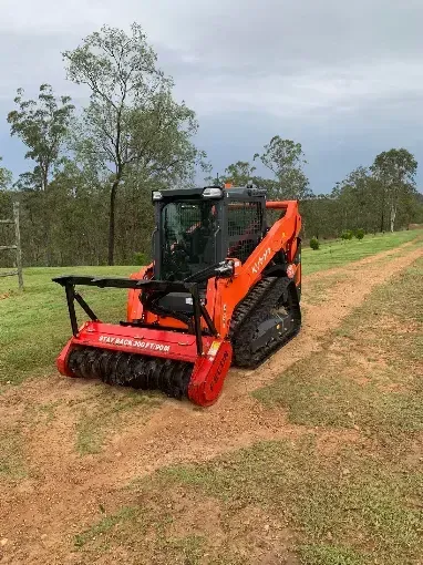 Orange skid steer with mower attachment clearing grass on a dirt track in a rural field