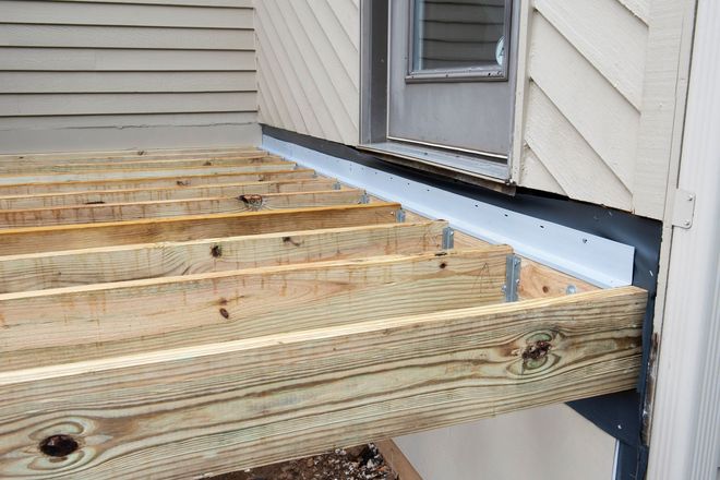 Wooden deck framing under construction beside a house, with joists and ledger board visible.