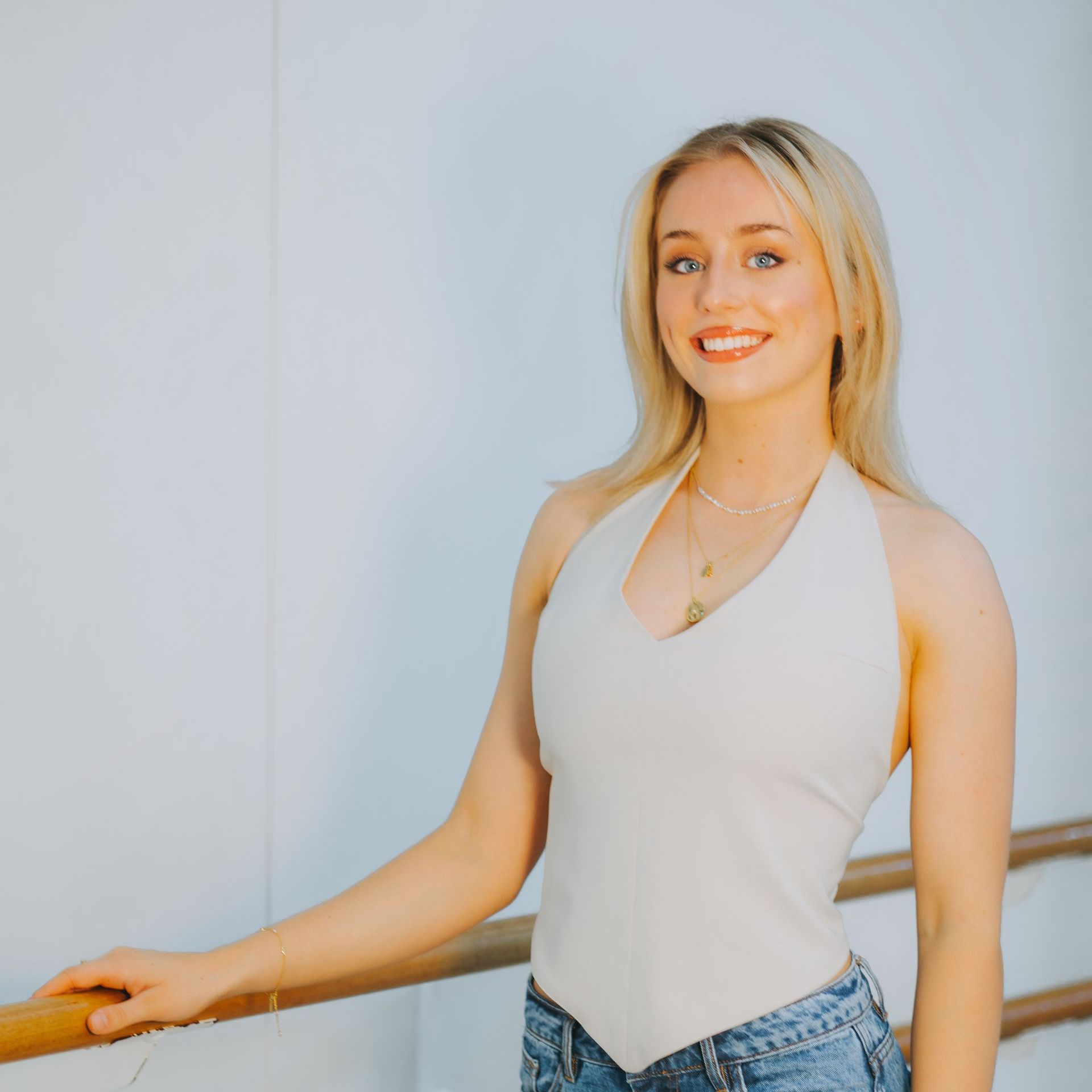 A woman in a white tank top and blue jeans is standing next to a railing and smiling.