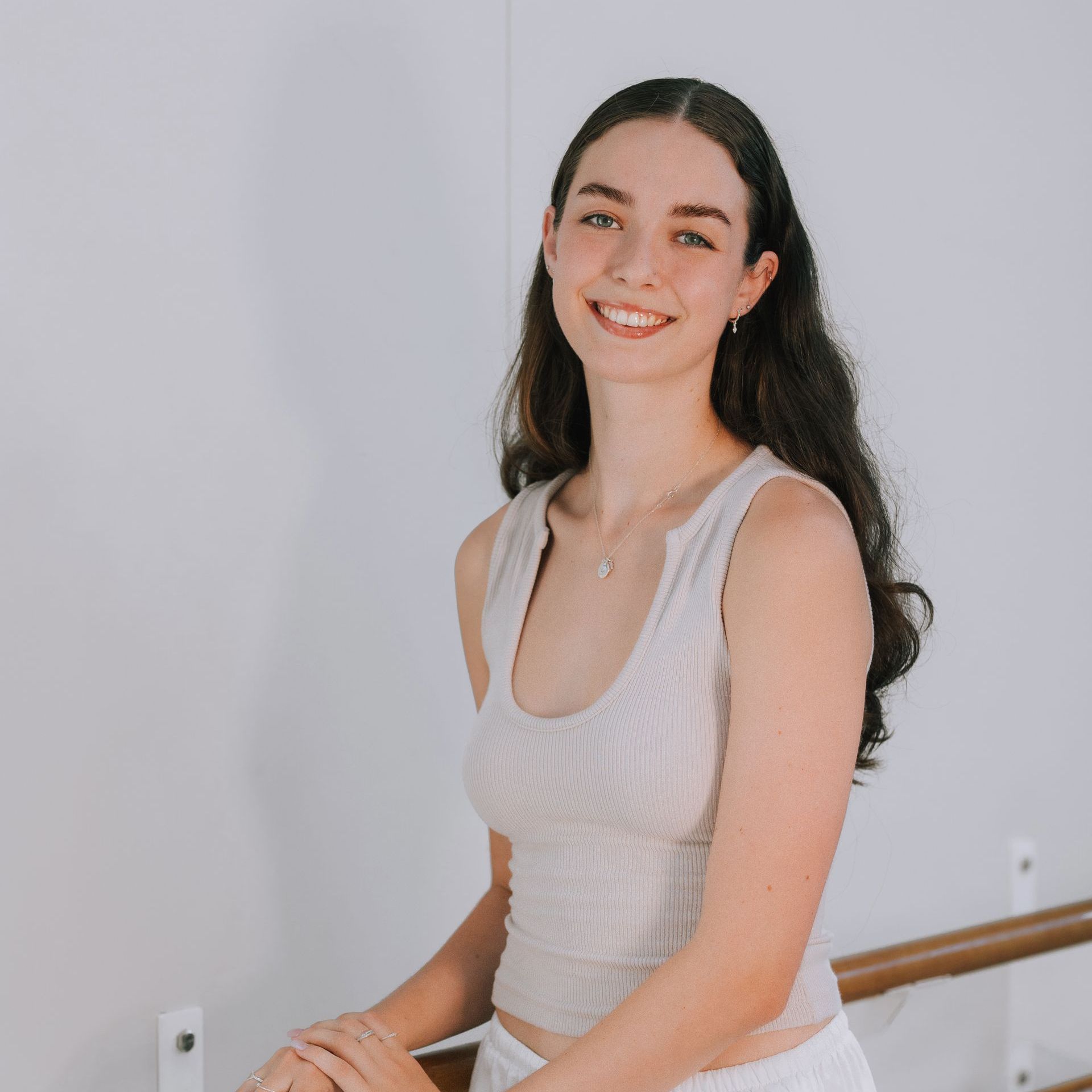 A woman in a white tank top is smiling while sitting on a bed.