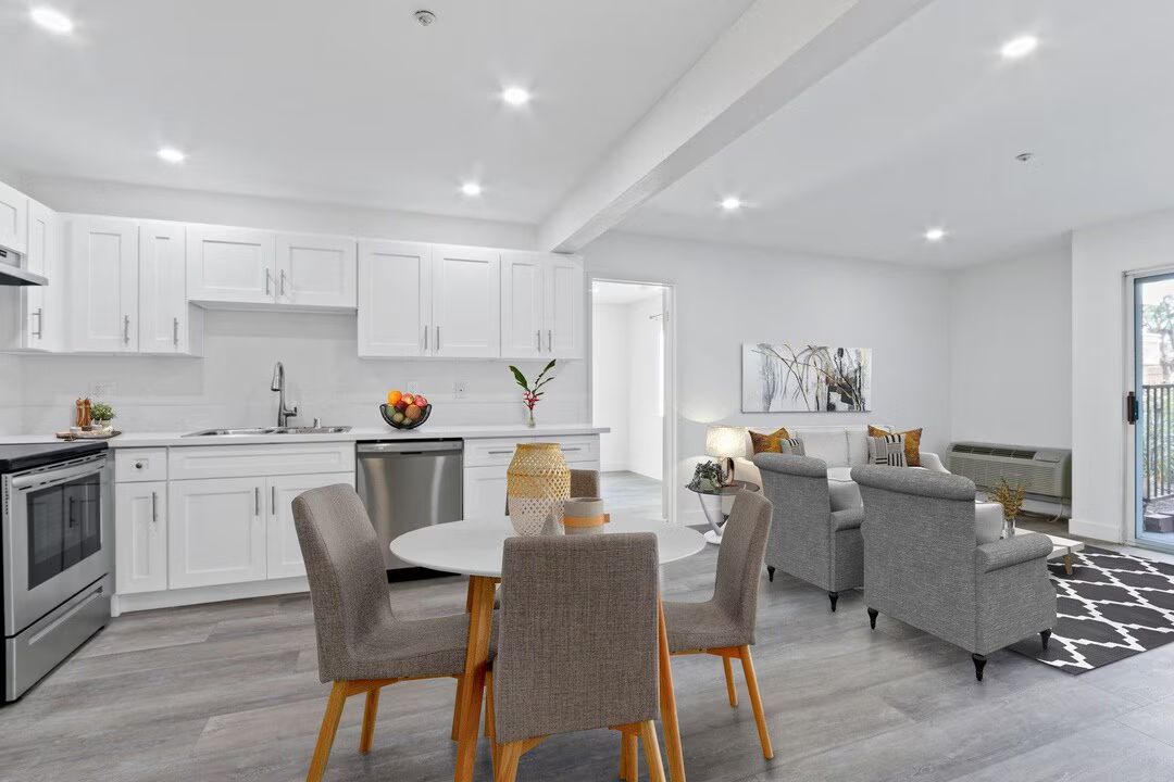 Bright open kitchen and living room with white cabinets, gray chairs, and gray sofa on a patterned rug