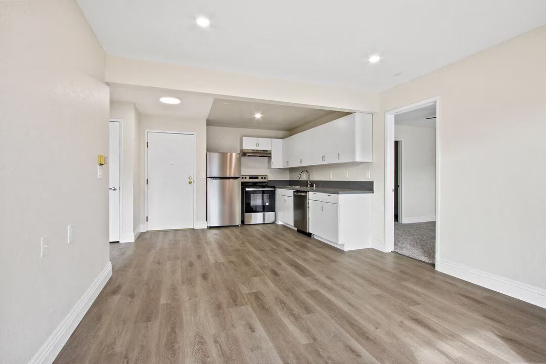 Empty modern apartment living area with wood floors, white walls, and open kitchen.