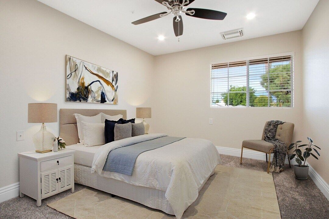 Bright bedroom with a gray bed, white nightstands, ceiling fan, abstract art, and a window with blinds.