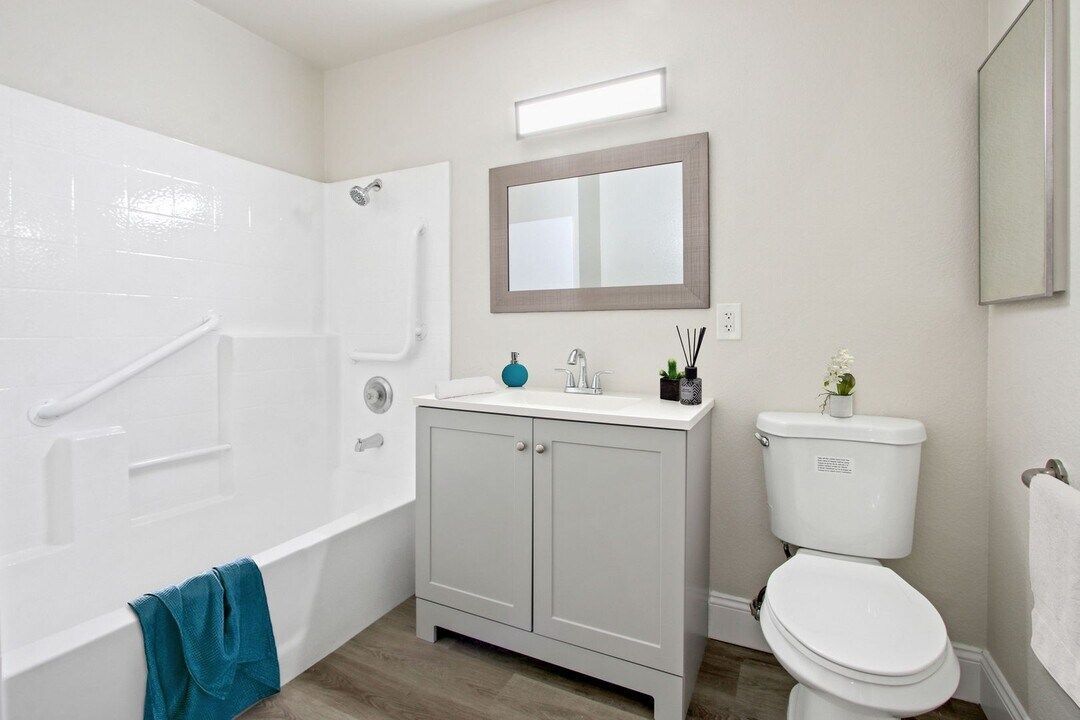 Bright bathroom with white tub, gray vanity, mirror, and toilet on wood-look floor.