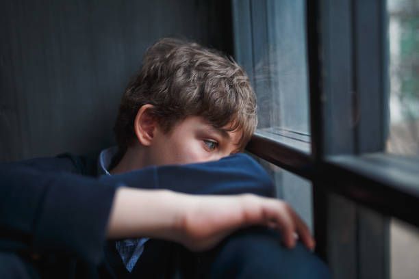 A young boy is sitting in front of a window looking out.