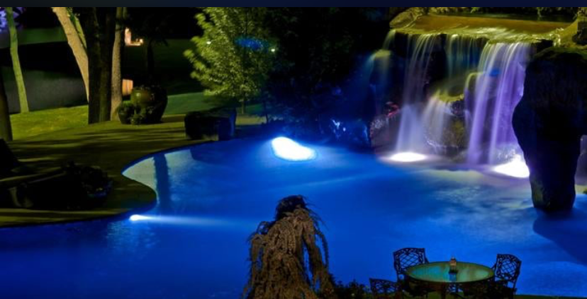 A swimming pool at night illuminated by blue lights, featuring a stone waterfall with cascading water and patio furniture.