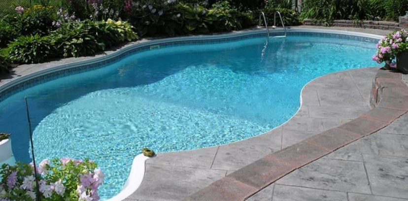 A backyard swimming pool with a free-form shape and stamped concrete patio, surrounded by lush green garden plants.