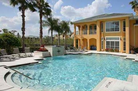 A bright turquoise swimming pool surrounded by a patio with palm trees and a multi-story yellow building in the background.