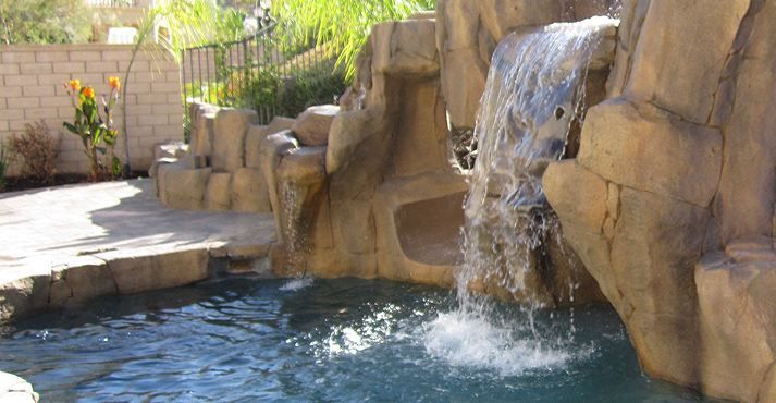 A decorative rock waterfall flows into a swimming pool in a sunny outdoor residential setting.