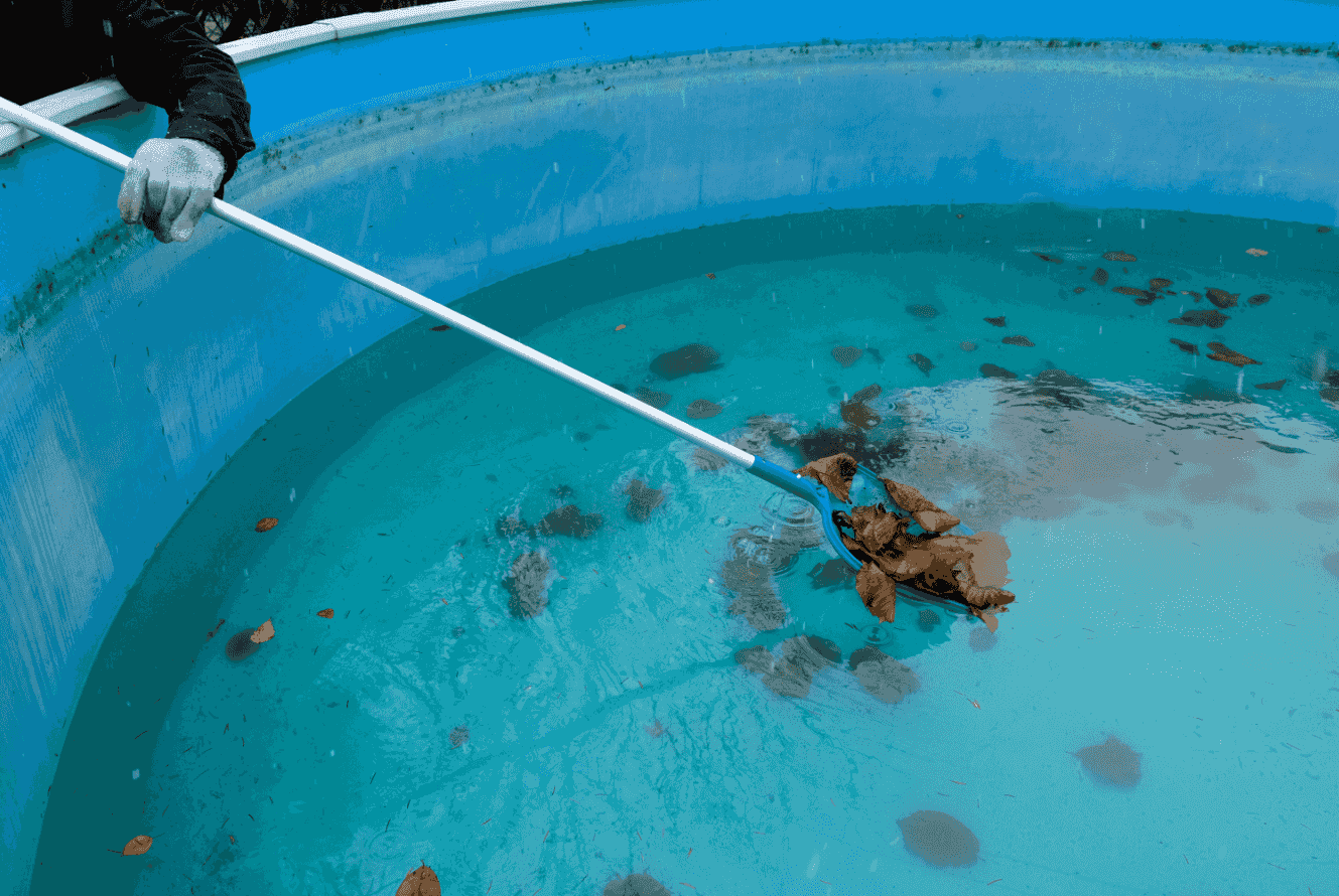 A person in a glove uses a long-handled net to remove debris from a bright blue, circular pool.