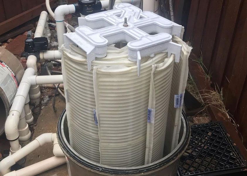 A dirty swimming pool DE filter grid assembly pulled out of its housing for cleaning, with PVC pipes in the background.