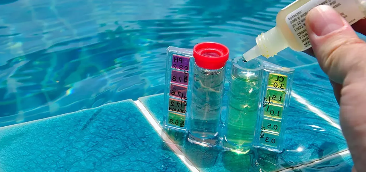 A hand adds drops to a pool water testing kit with color-coded scales beside a pool edge.