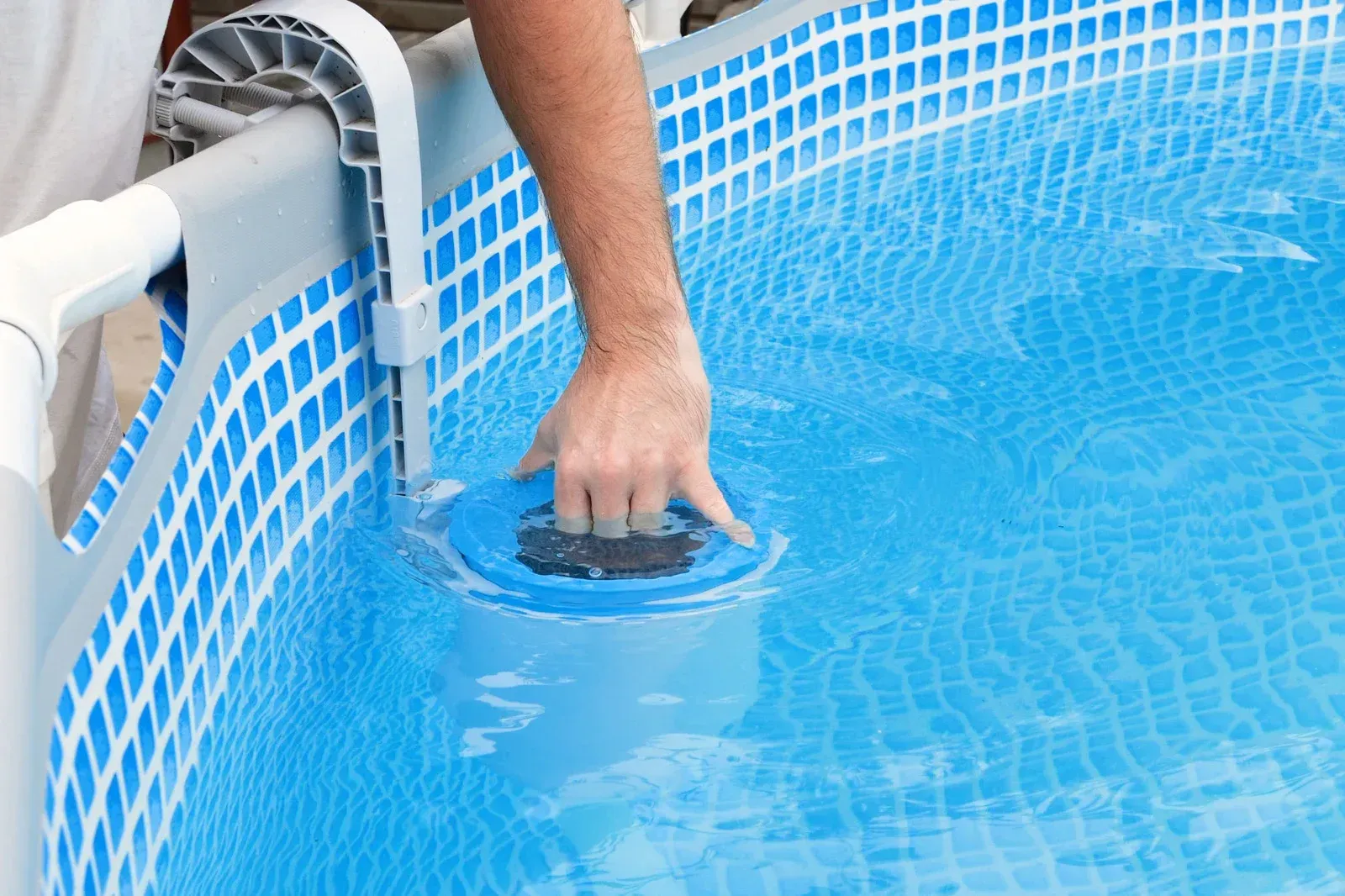 A person's hand reaches into a blue plastic pool to adjust a floating chlorine dispenser near the edge of the frame.