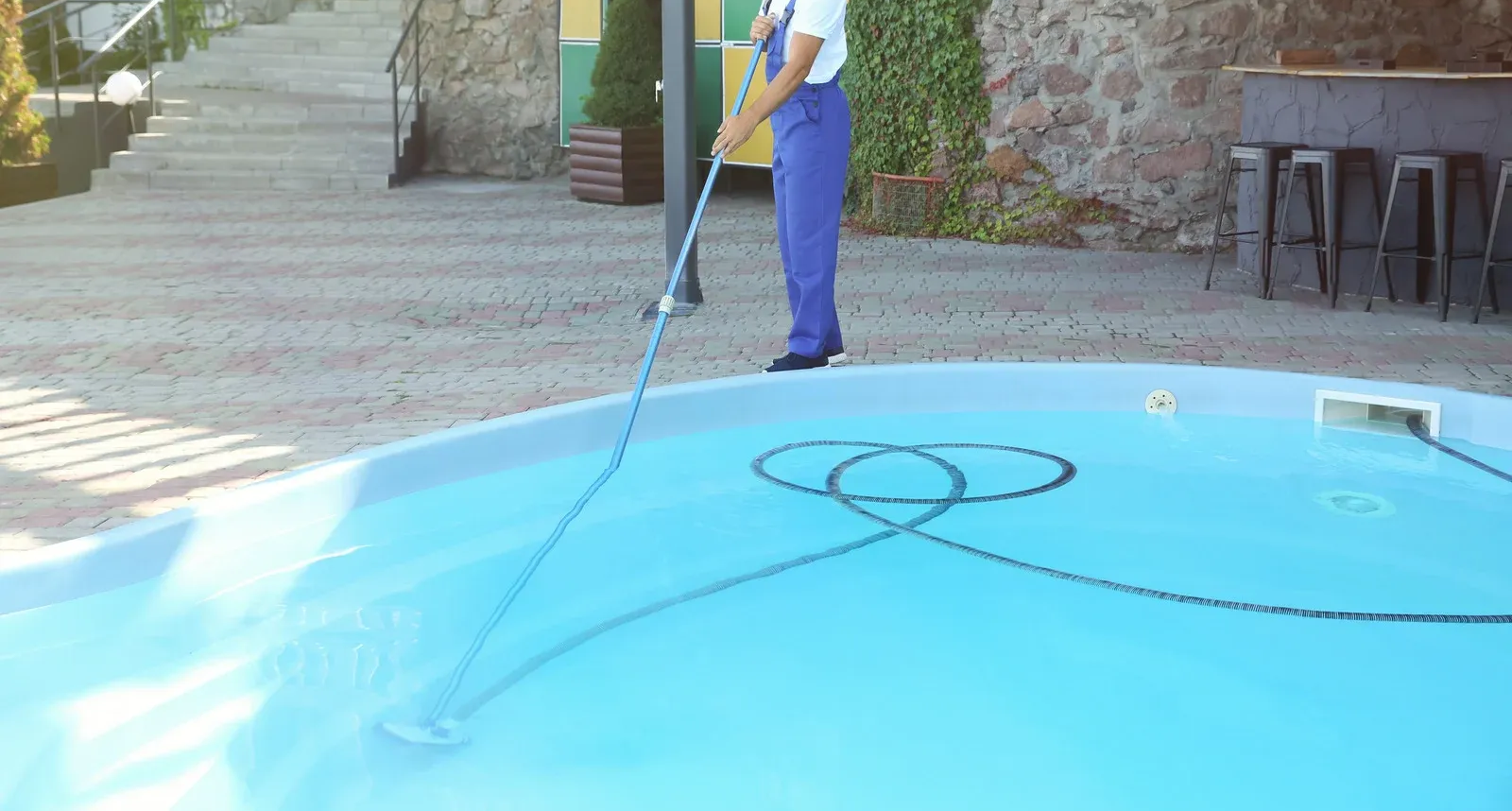 A person in blue overalls uses a long-handled brush to clean a round, blue swimming pool on a patio.