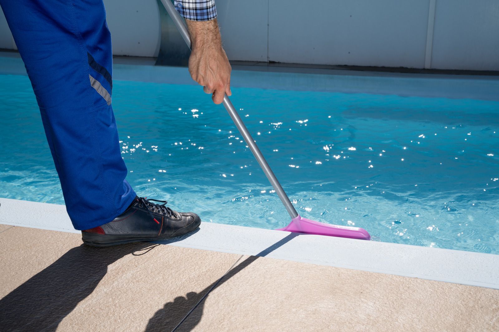 A person in blue work pants cleans a swimming pool edge with a long-handled brush.