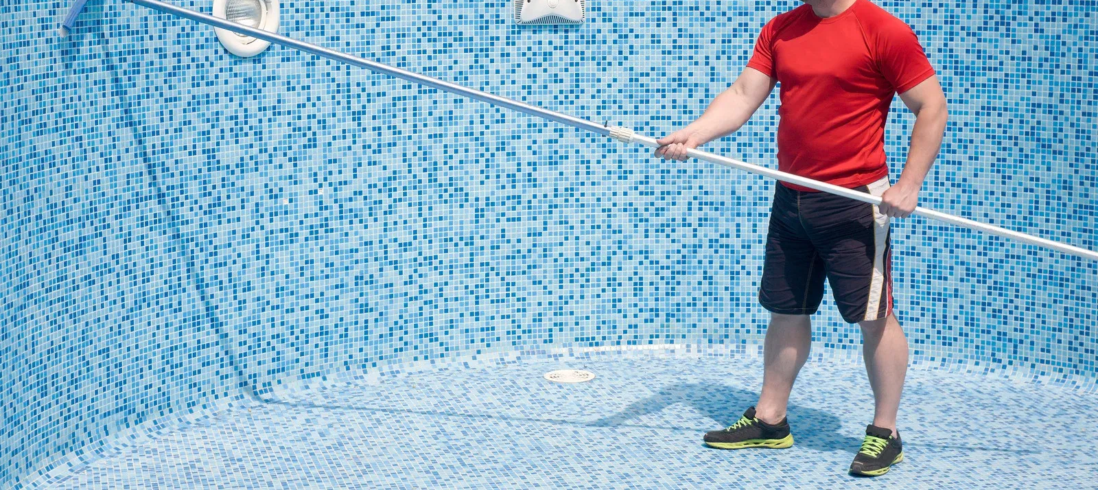 A person in a red shirt and black shorts stands in an empty, blue-patterned pool using a cleaning pole.