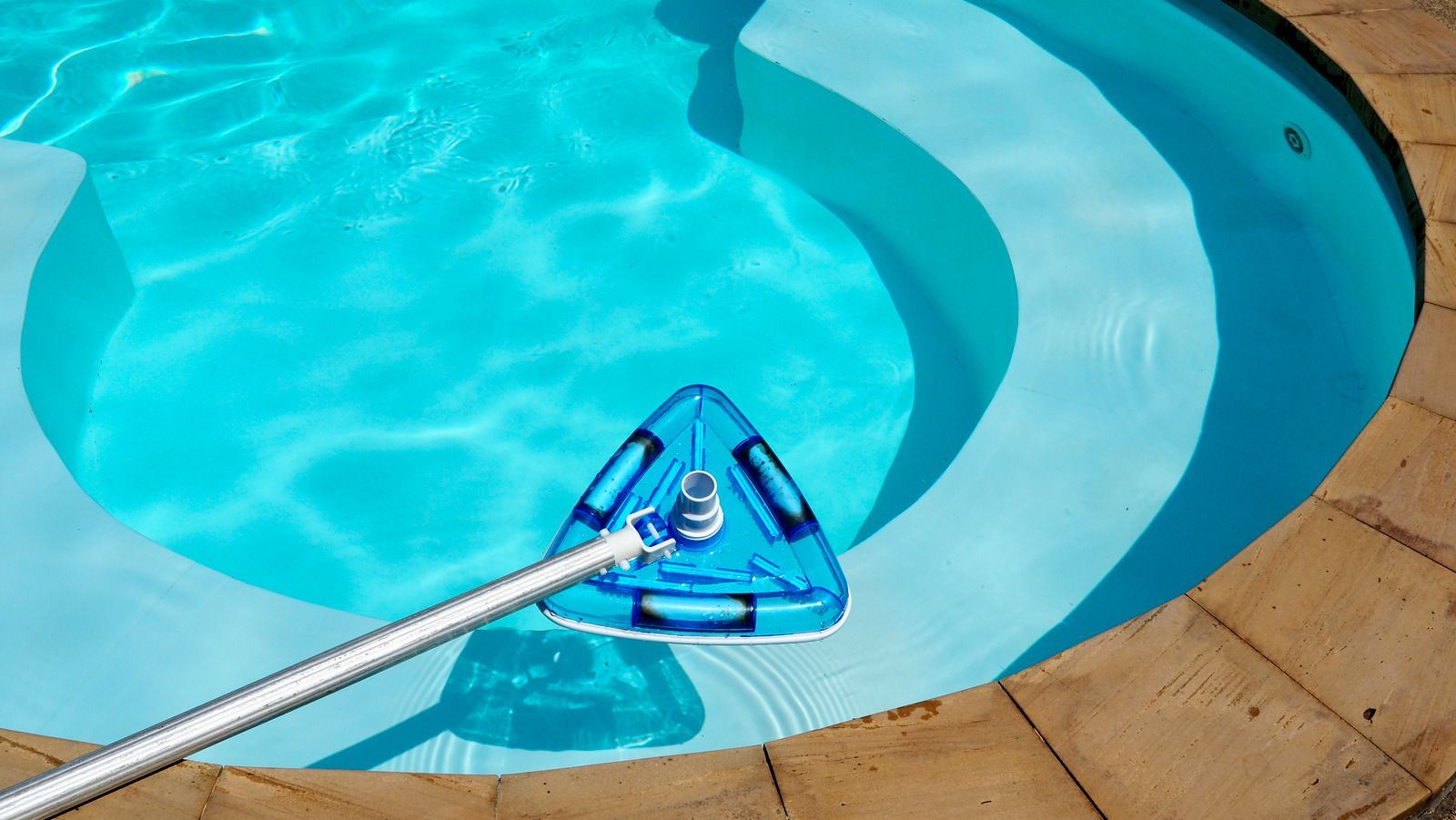 A triangular blue pool vacuum head attached to a long pole cleans the steps of a light blue swimming pool.