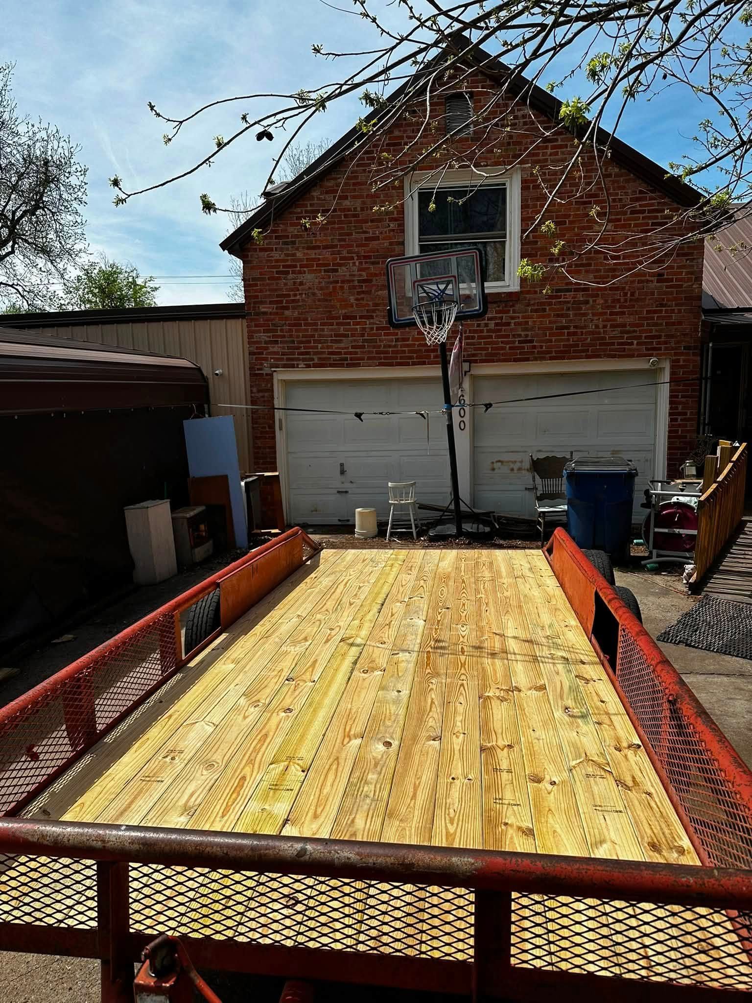 Red utility trailer loaded with plywood, parked in front of a brick garage with a basketball hoop.