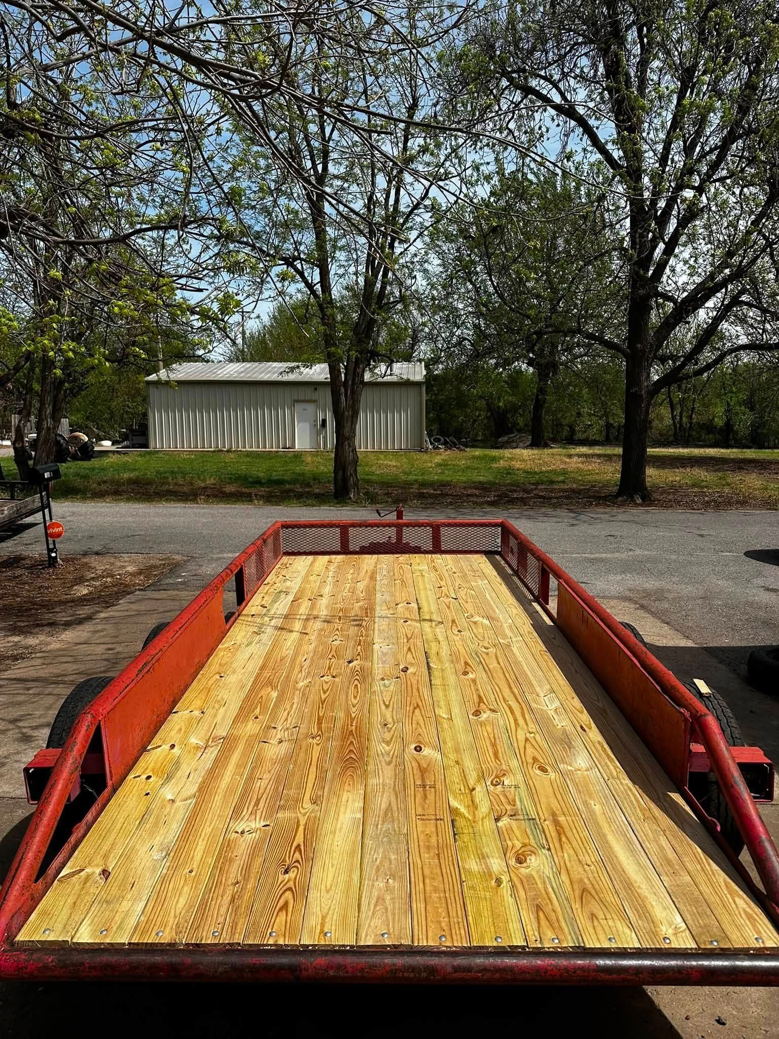 Red flatbed trailer with wooden deck on a gravel lot beneath trees and a small white shed.