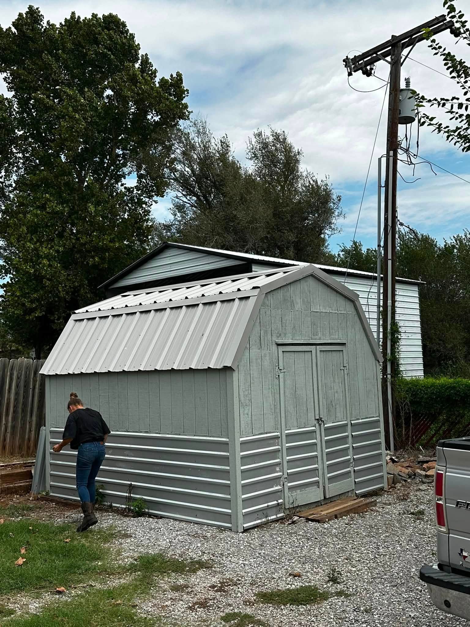 Small gray shed with a person standing beside it in a gravel yard, trees and utility pole in the background