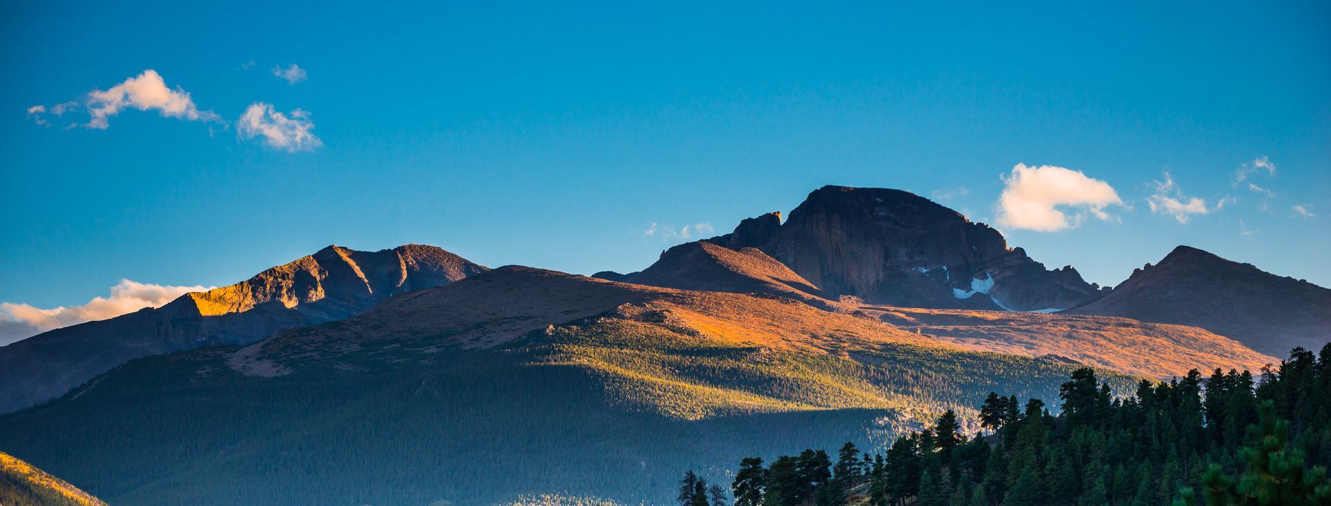 Sunlit mountain range under a blue sky with clouds, above a dark forest silhouette