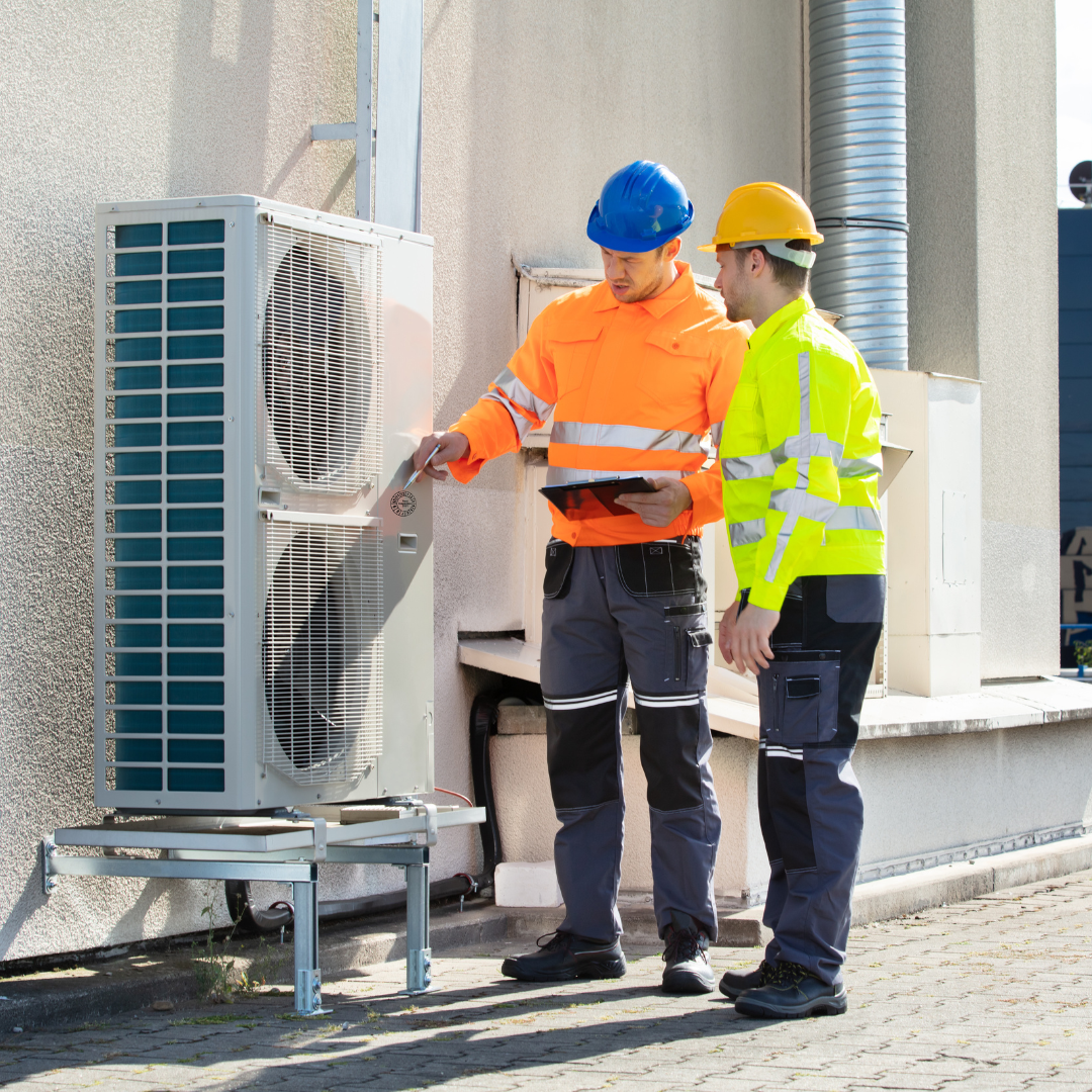Two men wearing hard hats are standing next to a large air conditioner.