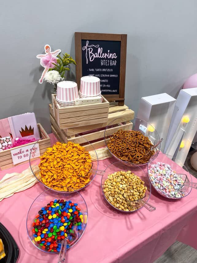 Pink-themed snack table with various treats in clear bowls, including candy and pretzels. A sign reads 