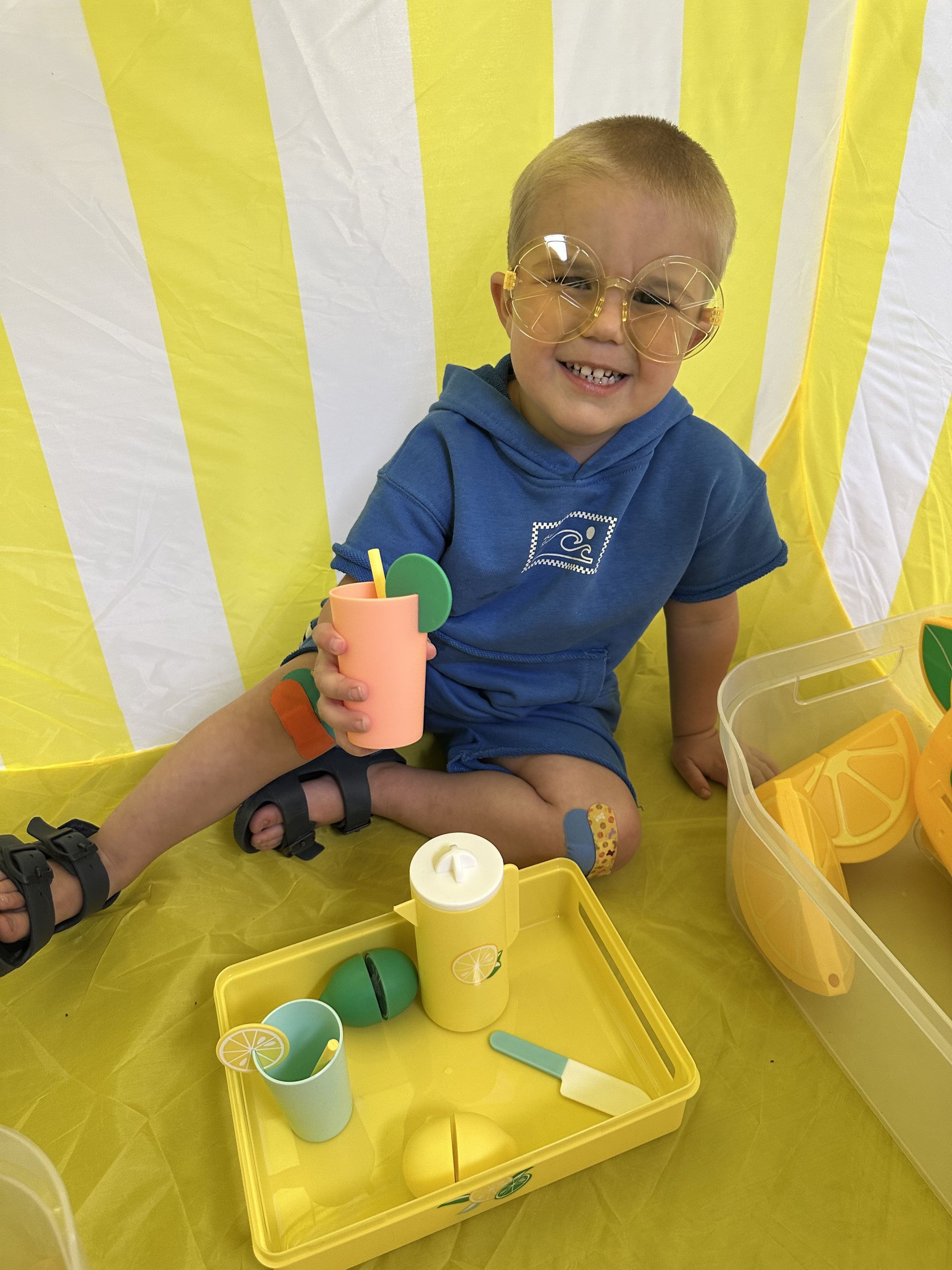 Young child with a toy drink, smiling, inside a yellow tent with toy fruits.