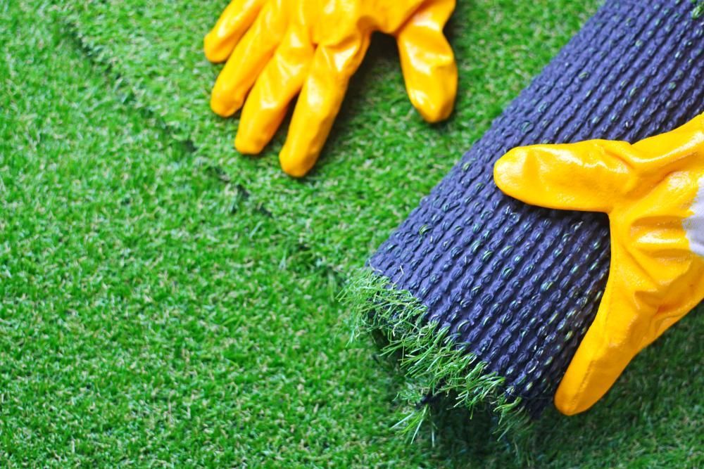 A Person Wearing Yellow Gloves Is Laying A Roll Of Artificial Grass On The Ground — Southern Bricks & Pavers In Moruya, NSW