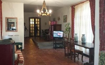Un soggiorno rustico con pavimento piastrellato, un lampadario, un televisore su un supporto, un tavolo di legno e tende rosse.