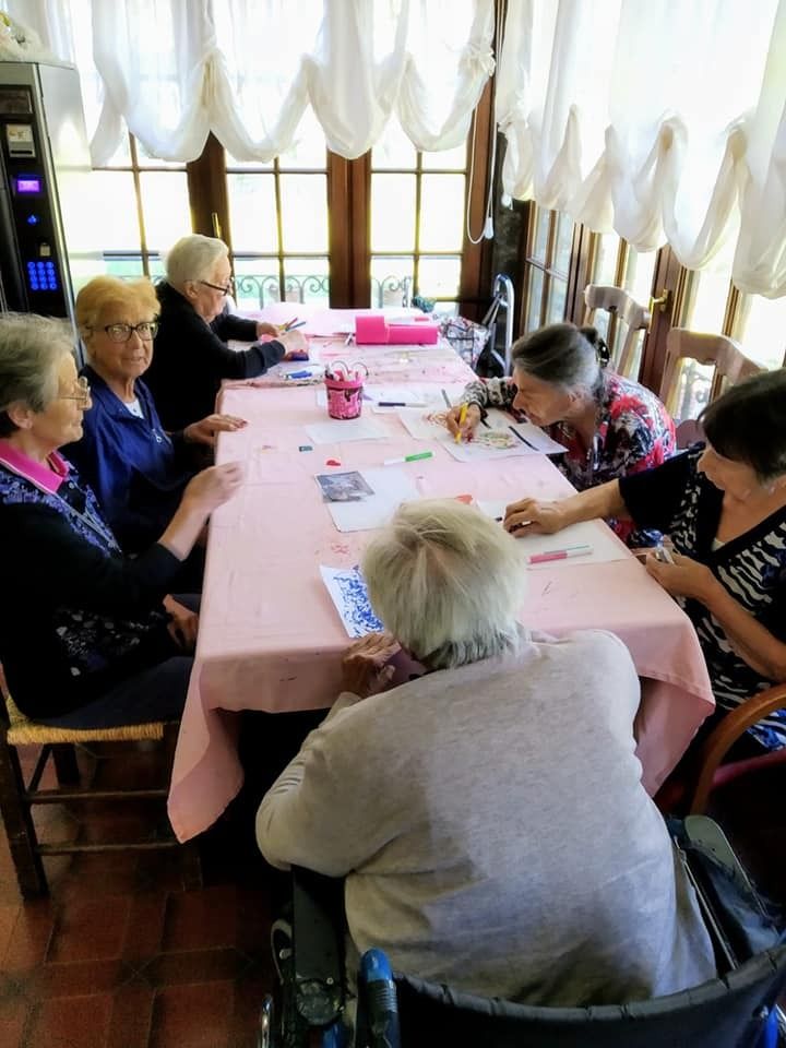 In una stanza illuminata dal sole, sei persone siedono attorno a un lungo tavolo rosa chiaro, intente a dedicarsi a progetti artistici e artigianali.