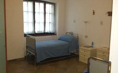 In una stanza spoglia, un singolo letto in stile ospedaliero con una coperta blu, posizionato accanto a una finestra e due comodini.