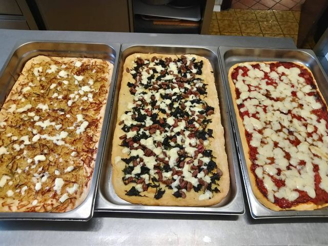 Su un bancone di metallo sono disposte tre teglie rettangolari di pizza, una con patate, spinaci e salsiccia e l'altra con la classica margherita.
